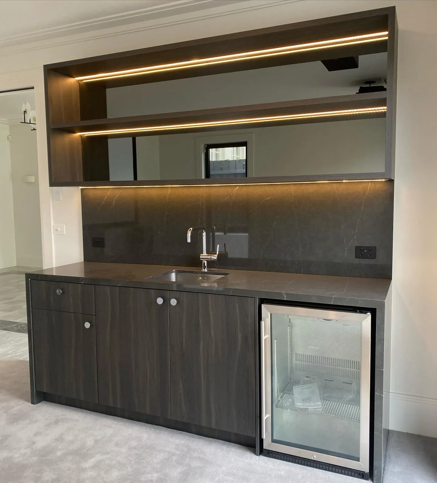 Dark oak wood-grain bar cabinetry with mirrored splashback and integrated bar fridge